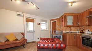 A compact living space with a sofa, a red leather ottoman, wooden cabinets, a kitchen area, and a door leading to another room. - The Aqua Shack (Newquay)