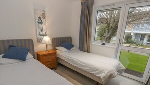 A bedroom with two single beds, a nightstand with a lamp, a window with curtains, and a view of a garden. - Pierpont D1 (Yarmouth)