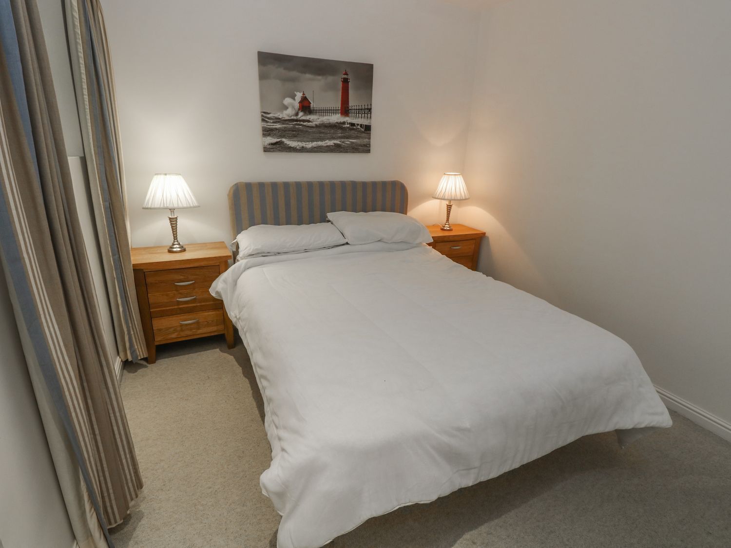 A neatly made bed with white linens, two bedside tables with lamps, a striped headboard, and a painting of a lighthouse on the wall.