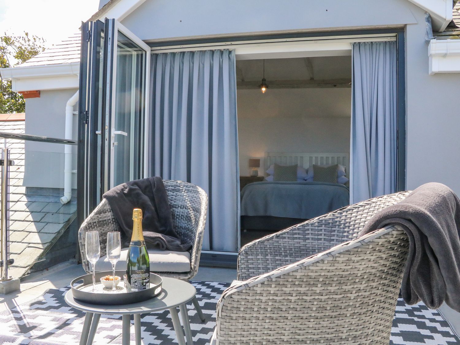 Un balcon avec un fauteuil en osier, une petite table avec une bouteille de champagne et des verres, et un lit visible à travers la porte coulissante ouverte.