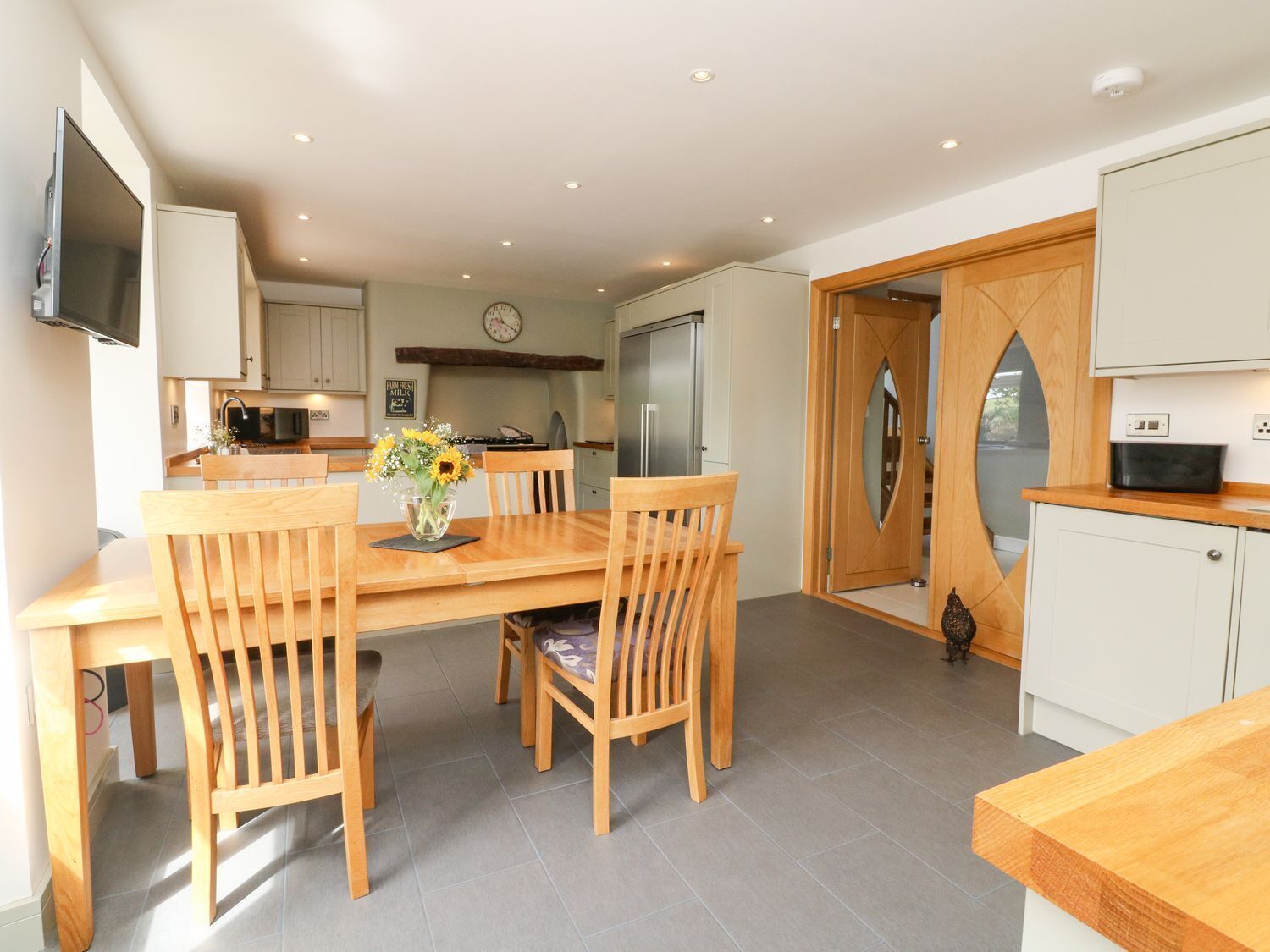 A modern kitchen with a dining area, featuring a wooden table and chairs, a clock on the wall, and a TV mounted above the counter.