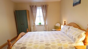 A bedroom with a bed, wooden headboard, a nightstand, a dresser, a window with curtains, and a painting on the wall. - Portstewarts Home From Home (Portstewart)