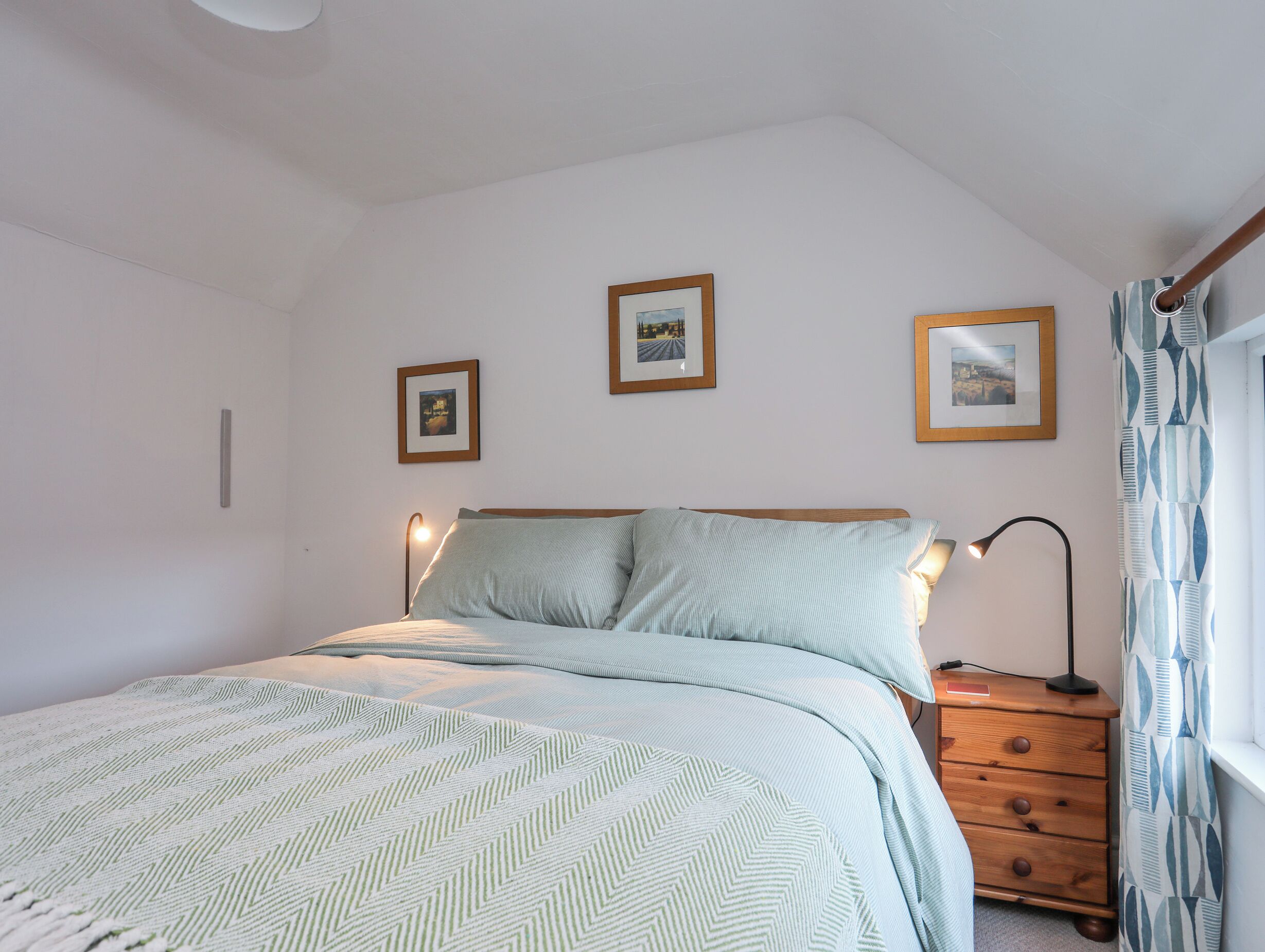 A bedroom with a bed, two bedside lamps, a nightstand, and three framed pictures on the wall.