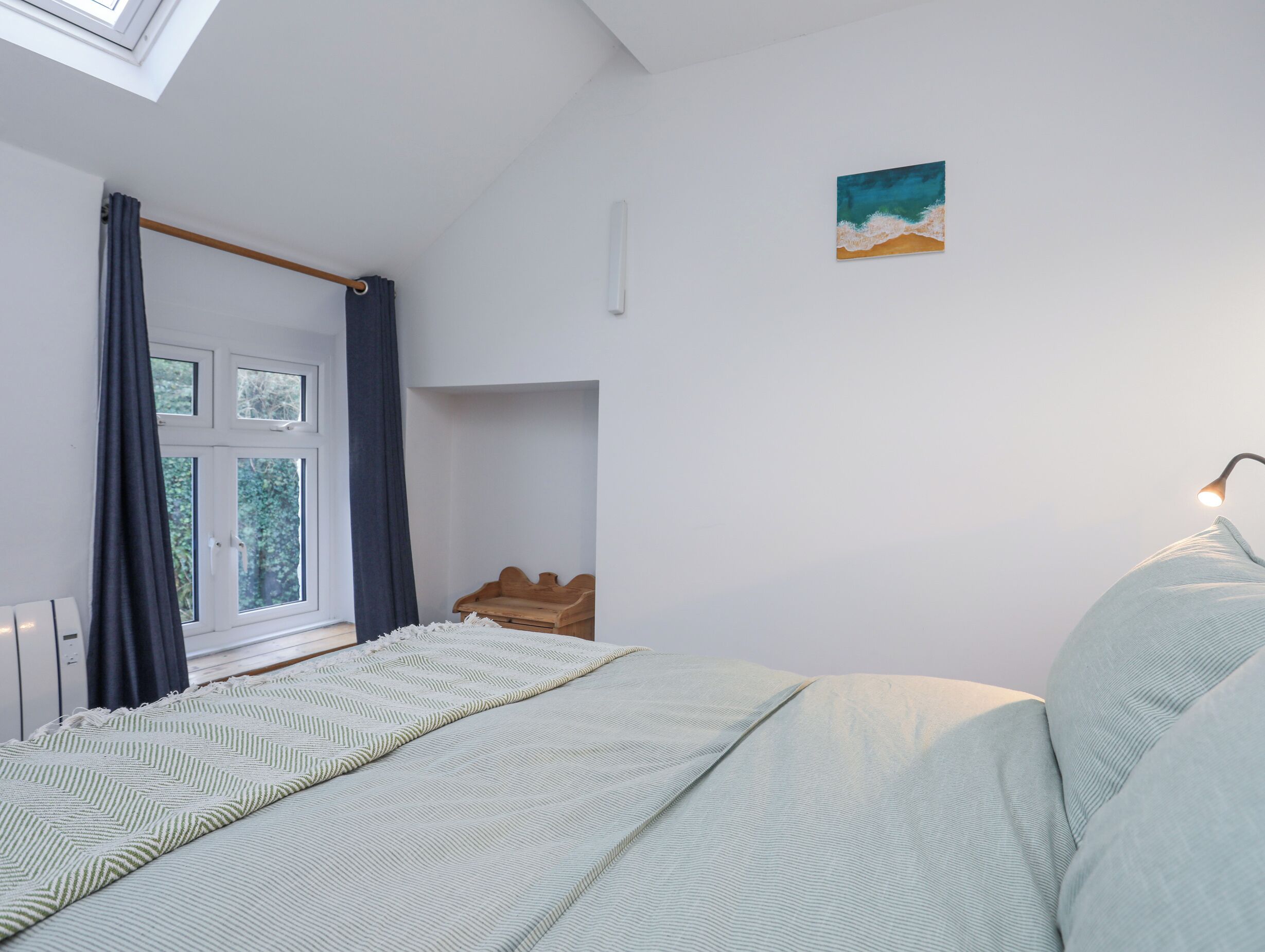 A bedroom with a bed, a window with curtains, a painting, and a skylight.