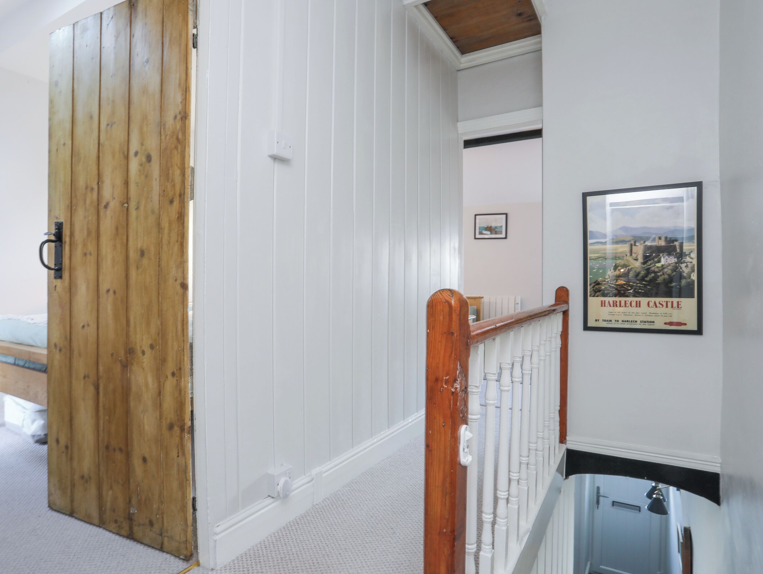 A hallway with a wooden door, white walls, a white railing, and a poster of Harlech Castle.