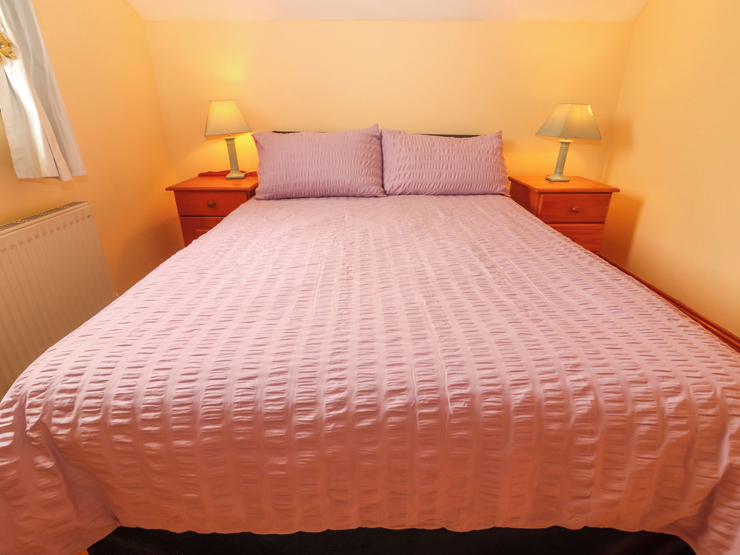 A neatly made bed with a pink bedspread and two pillows, flanked by two bedside tables with lamps.