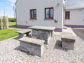 A building with a white exterior, two windows with purple frames, and a stone table with benches in front.