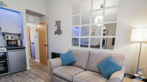 A compact living space with a kitchenette, a grey sofa with blue cushions, a patterned rug, and a decorative wall piece. - Estuary Escape (Porthmadog)