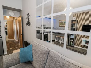 A modern living room with a grey sofa and a blue pillow, a kitchen with white cabinets and a tiled backsplash, and a dining area with two wooden stools. - Estuary Escape (Porthmadog)