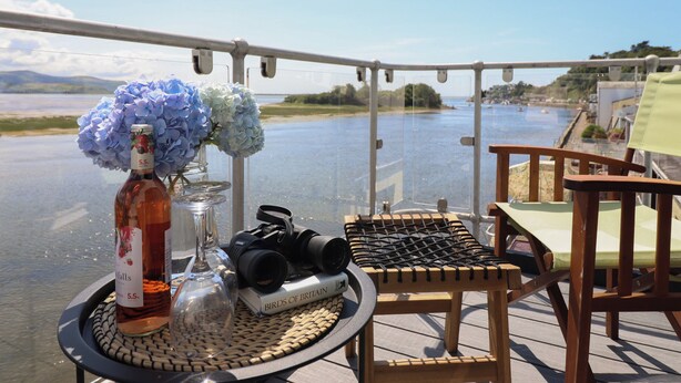 A table with a bottle of wine, a glass, a camera, and a book on a deck overlooking a body of water. - Estuary Escape (Porthmadog)