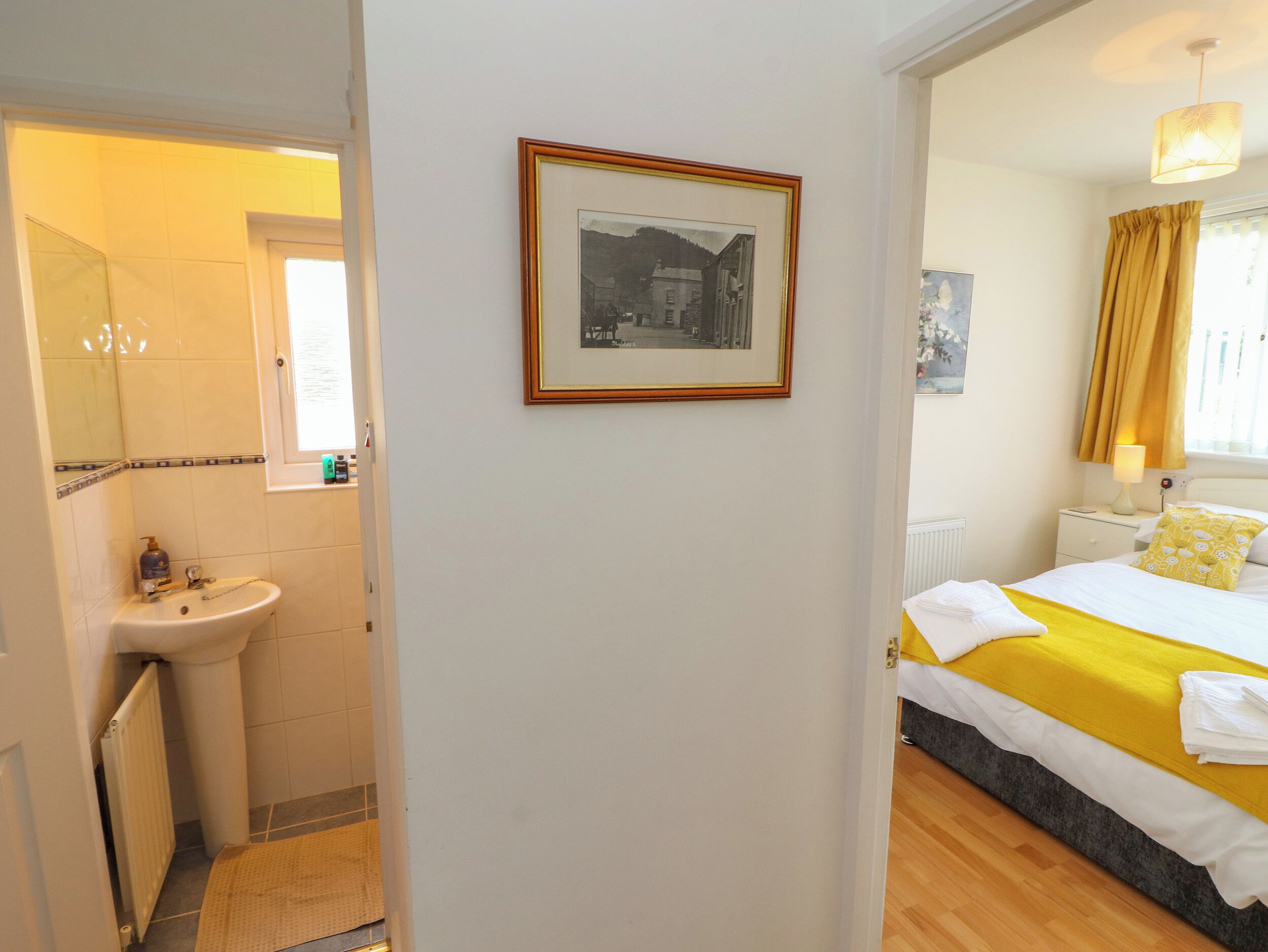 Ein Hotelzimmer mit Bett, Nachttisch, Lampe, einem gerahmten Bild an der Wand, Badezimmer mit Waschbecken und Spiegel sowie einem Fenster mit Vorhängen.