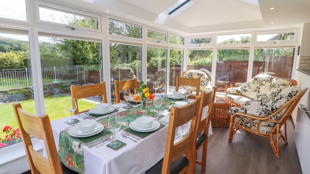 A conservatory with a dining table set for six, surrounded by wicker chairs and a sofa. - Craggy View (Kendal)