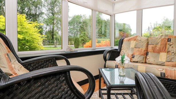 Une véranda meublée de mobilier en osier, avec une table en verre et une vue sur les arbres et les fleurs.