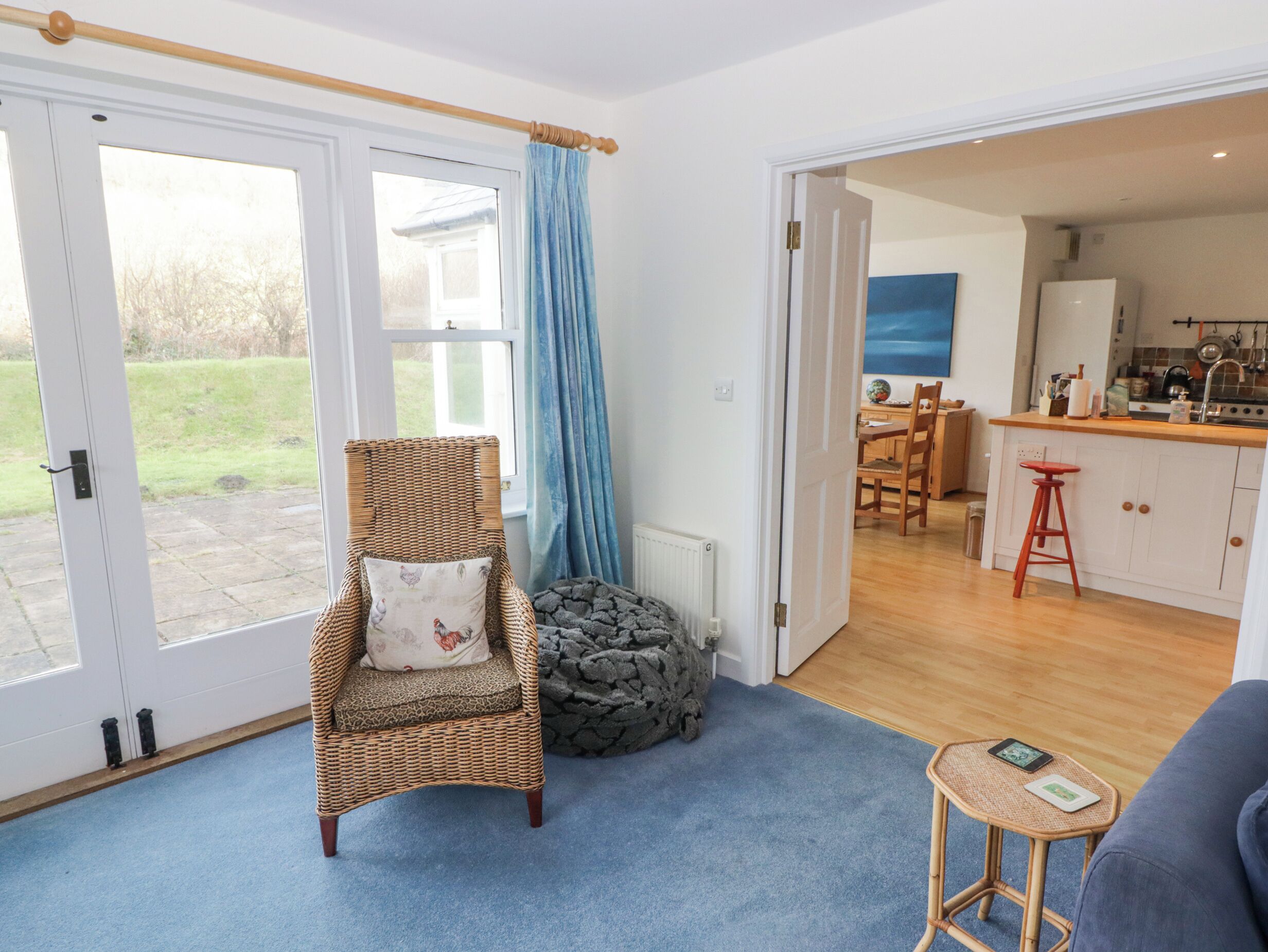 A room with a wicker chair, a small stool, a blue carpet, and a view of a kitchen and garden through a glass door.