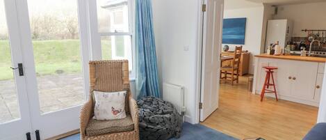 A room with a wicker chair, a small stool, a blue carpet, and a view of a kitchen and garden through a glass door.