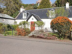 A white house with a red door, surrounded by a garden with various plants and a large flowering bush. - Fuchsia Cottage (Fortrose)