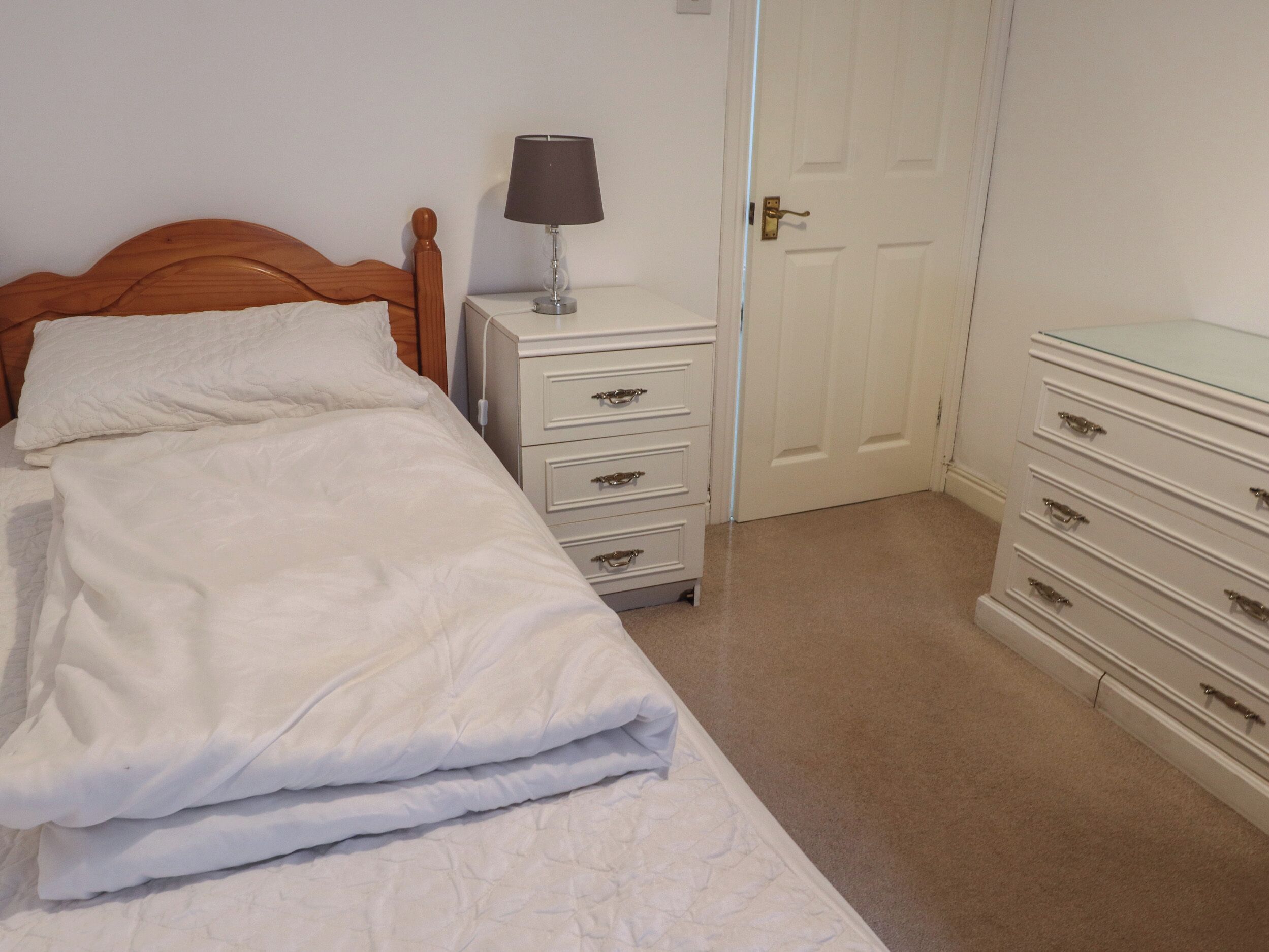 A neatly made bed with a white comforter and pillows, a bedside table with a lamp, and a dresser with drawers.