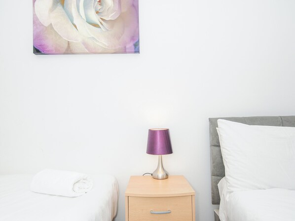 A neatly made bed with white linens, a wooden nightstand with a lamp, and a wall-mounted painting of a flower. - 24 Valley Lodge (Gunnislake)