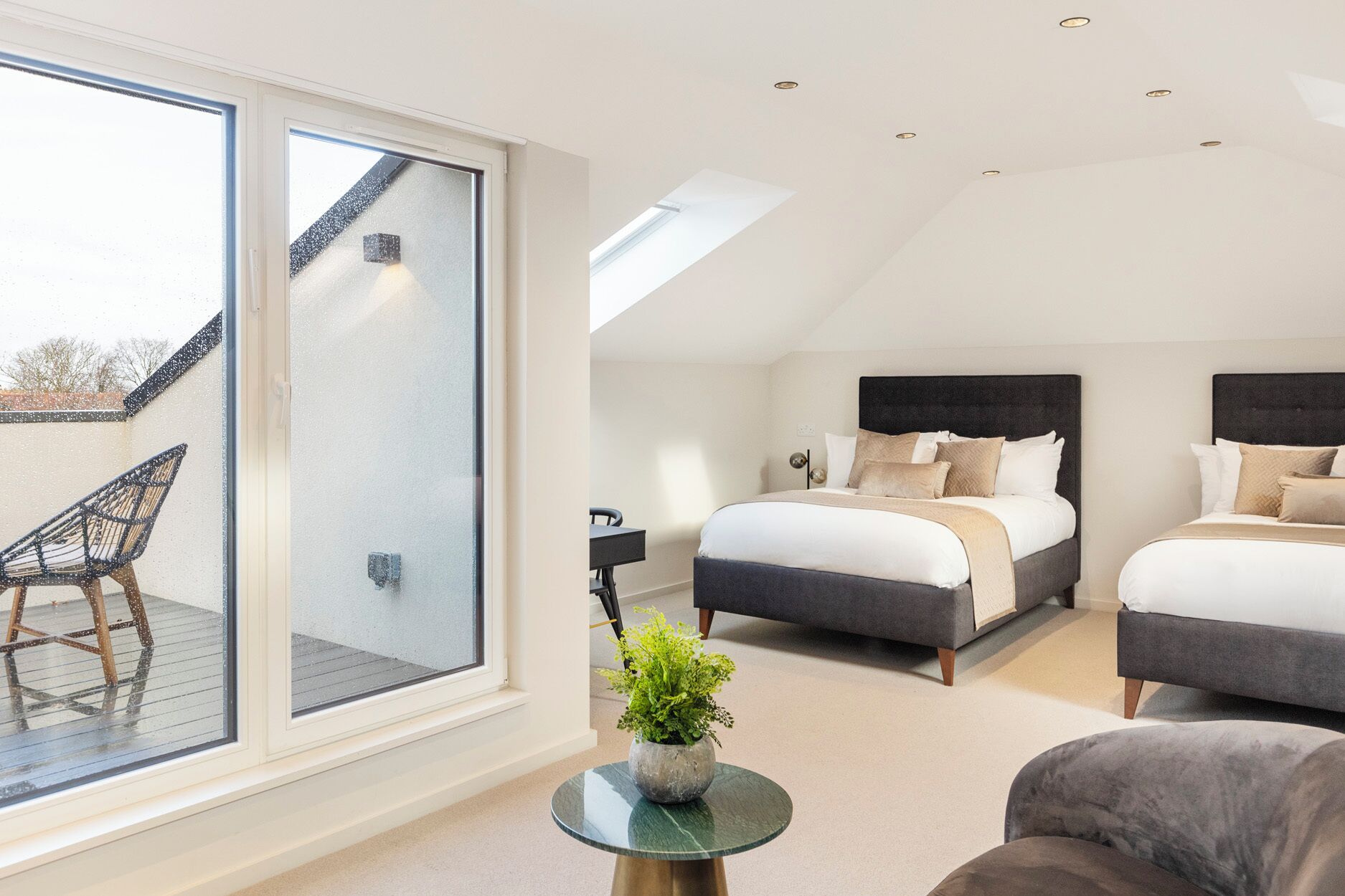 A modern bedroom with two beds, a large sliding glass door, and a small round table with a plant.