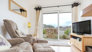 A modern living room with a sofa, a television, and a view of a cityscape through large windows. - 66D South Snowdon Wharf (Porthmadog)