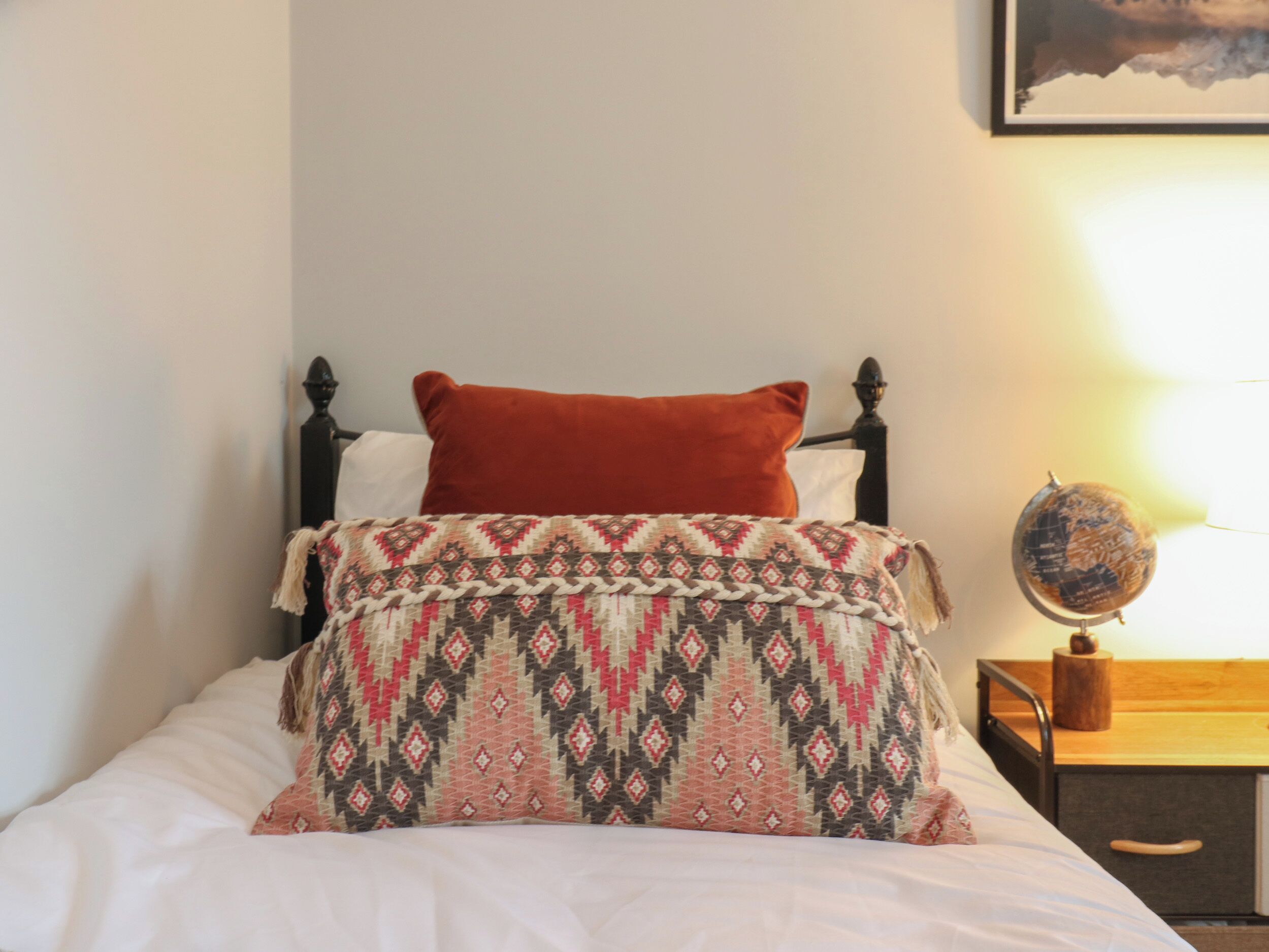 A bed with a patterned blanket, a red pillow, and a wooden nightstand with a globe and a wooden object.