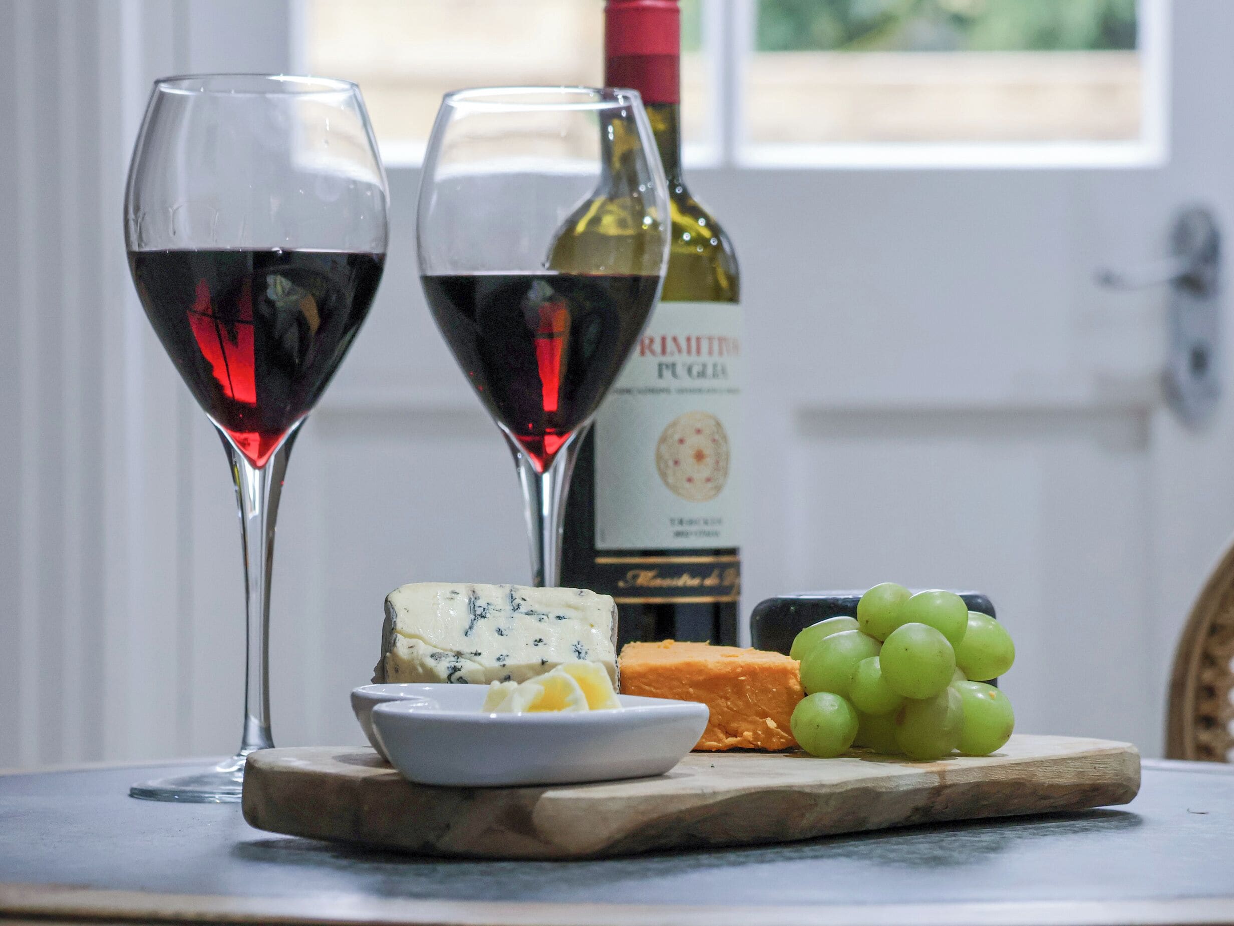 Dos copas de vino, una botella, un bloque de queso, un cuenco de mantequilla y un racimo de uvas sobre una tabla de madera.