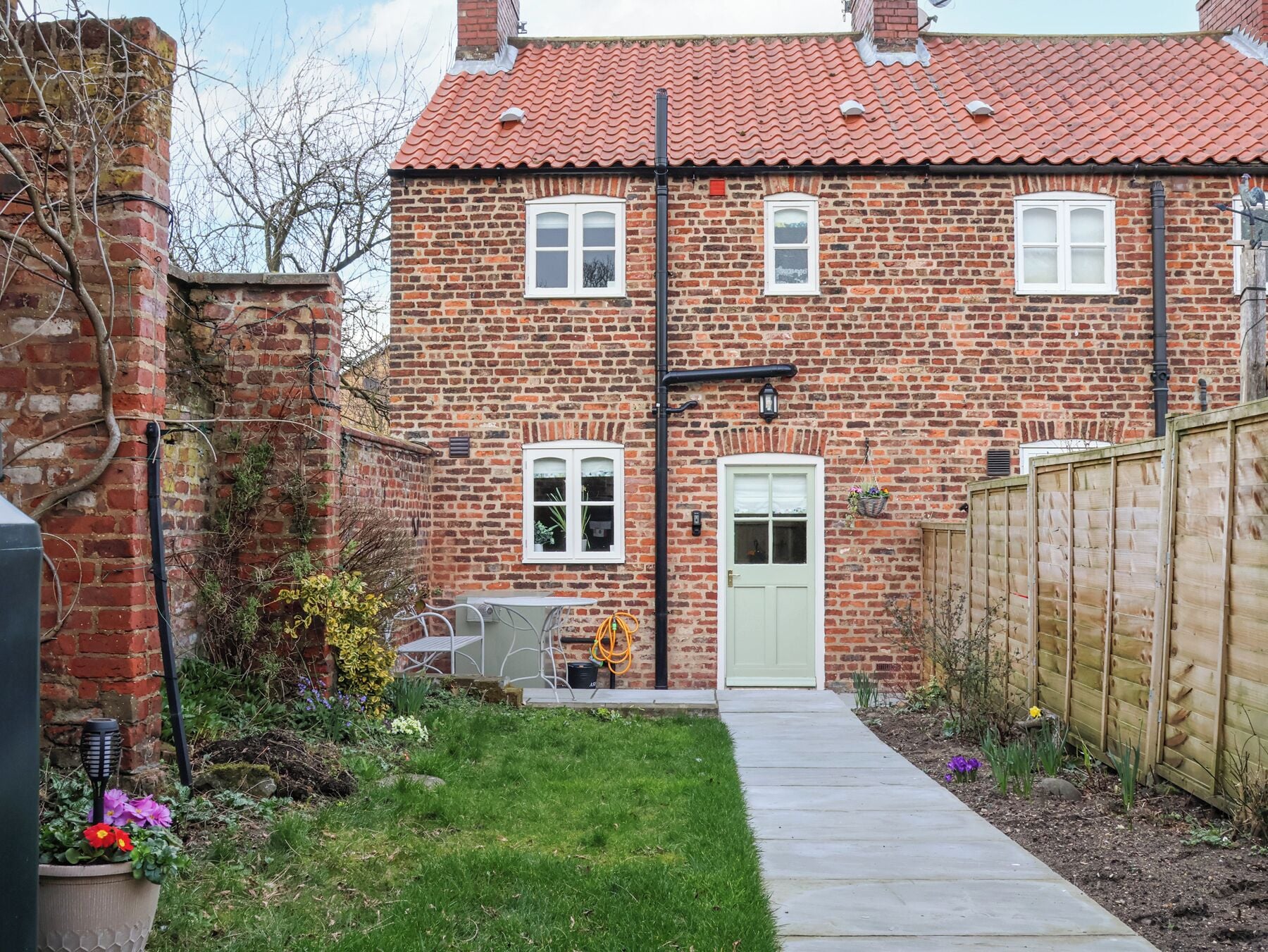 A two-story brick house with a red-tiled roof, a front door, and a small garden with flowers and a pathway leading to the door.