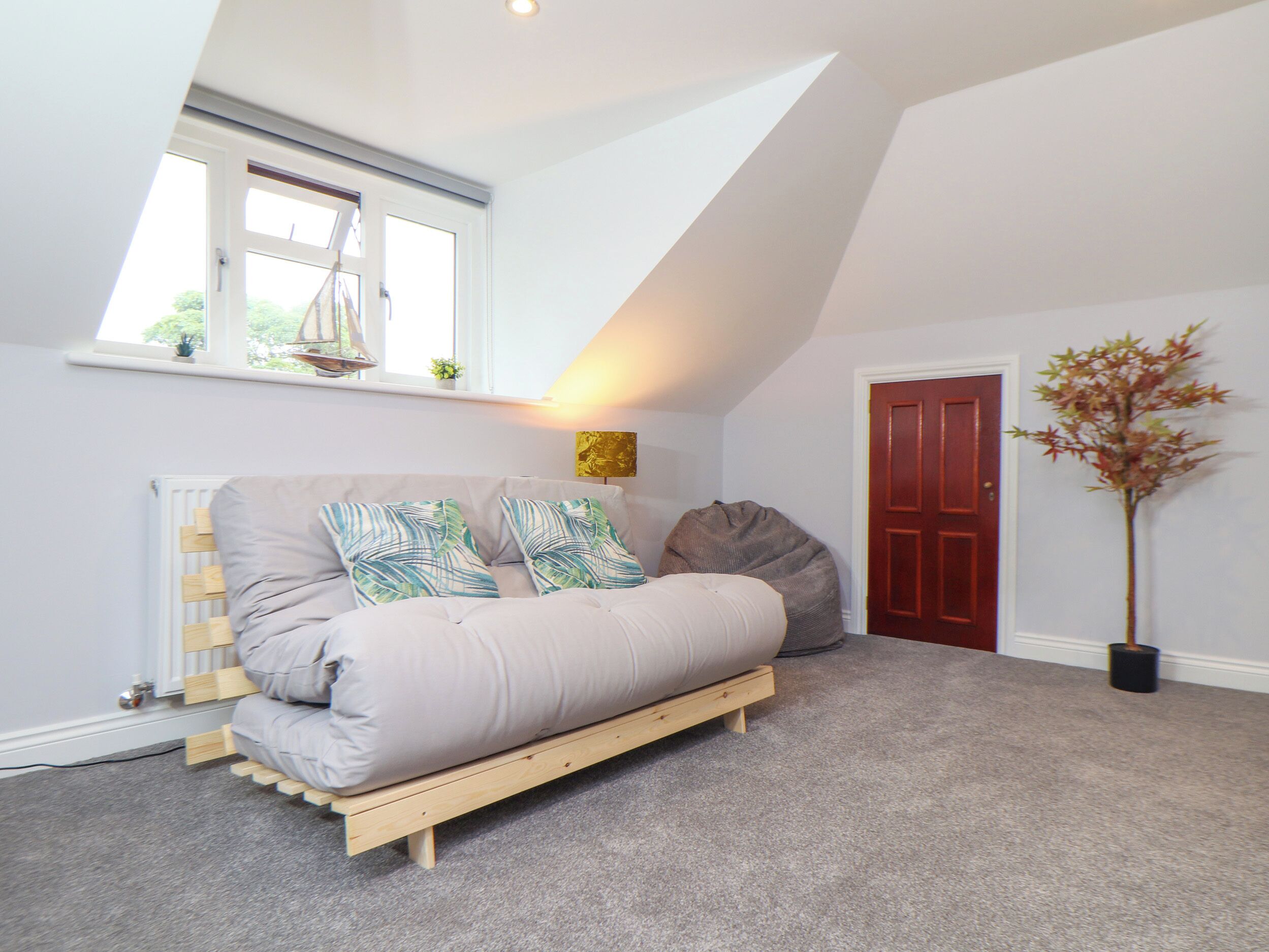 Une chambre de style « loft » avec un plafond en pente, un lit-banquette intégré en bois, un radiateur, une fenêtre avec vue, une porte et une plante en pot.