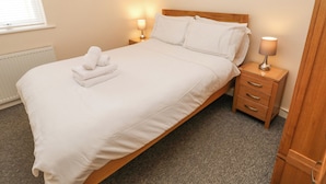 A neatly made bed with white linens and a folded towel on the footboard. There are two bedside lamps on wooden nightstands. A framed picture hangs on the wall above the bed.