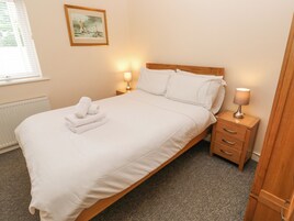 Cama bem arrumada com lençóis brancos e uma toalha dobrada na cabeceira. Duas luminárias de cabeceira estão sobre mesas de madeira. Uma moldura com uma fotografia está pendurada na parede acima da cama.