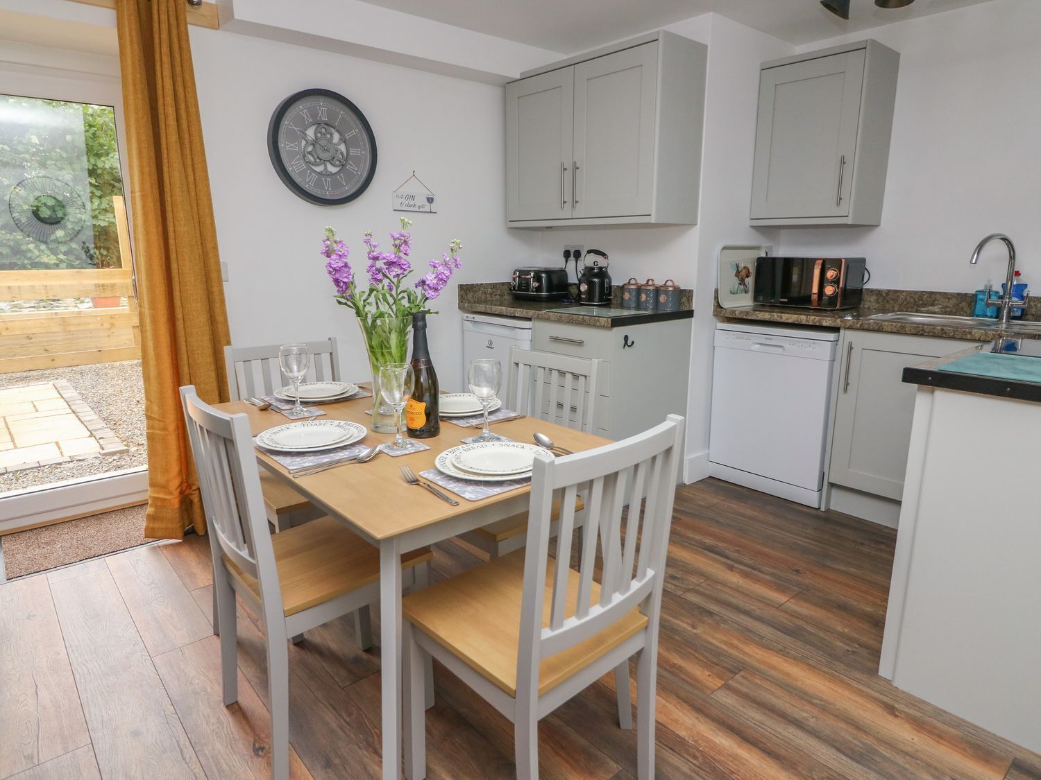 A modern kitchen with white cabinets, a wooden dining table set for four, and a clock on the wall.