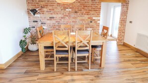 A dining area with a brick wall, wooden table and chairs, a hanging light fixture, and a potted plant. - Heron's Den (Horncastle)