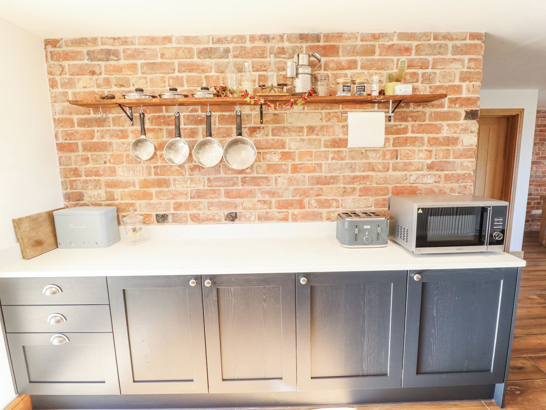 A kitchen with a brick wall, a wooden shelf with pots, a toaster, and a microwave.