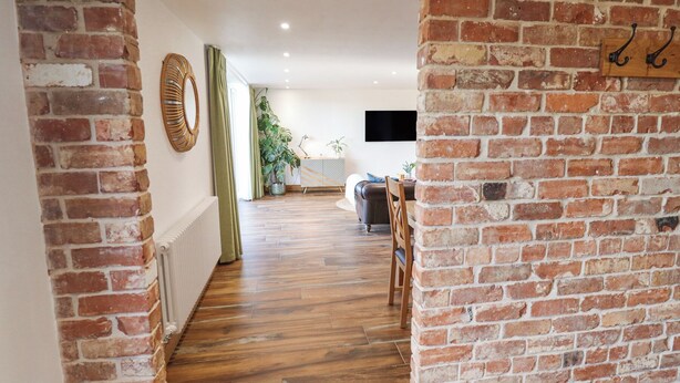A hallway with exposed brick walls, wooden flooring, and a view into a living area with a sofa and a TV. - Heron's Den (Horncastle)