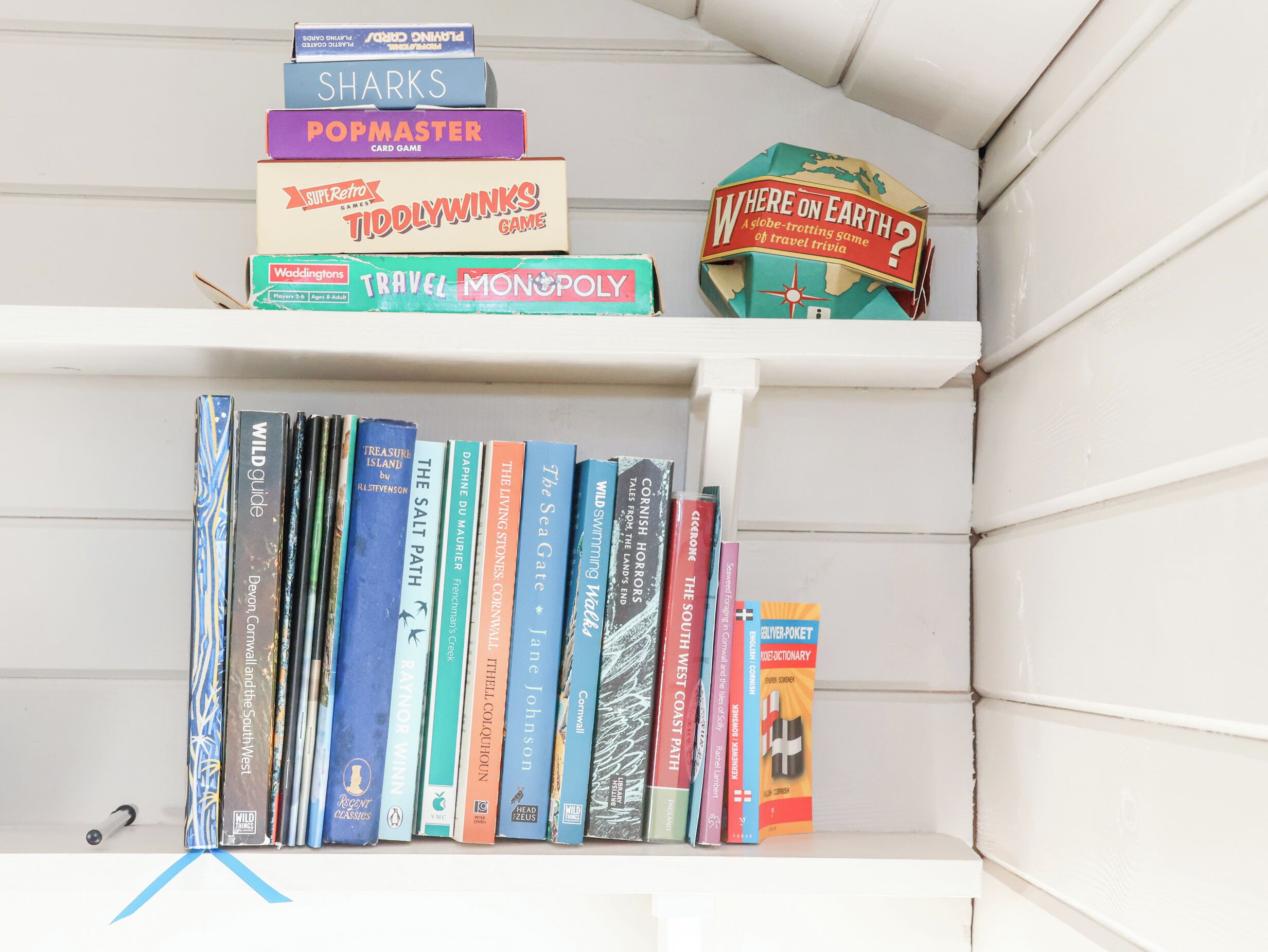 Une bibliothèque contenant divers livres et jeux, notamment « Sharks », « Popmaster », « Tidlywinks Game », « Where on Earth? », « Wild Guide », « Treasure Island », « The Salt Path », « The Living Stones », « The Sea Gate », « Cornish Horrors », « The South West Coast Path », « Wild Swimming Wales », « The South », « The Silver Picket » et « Dictionary of Cornish ».