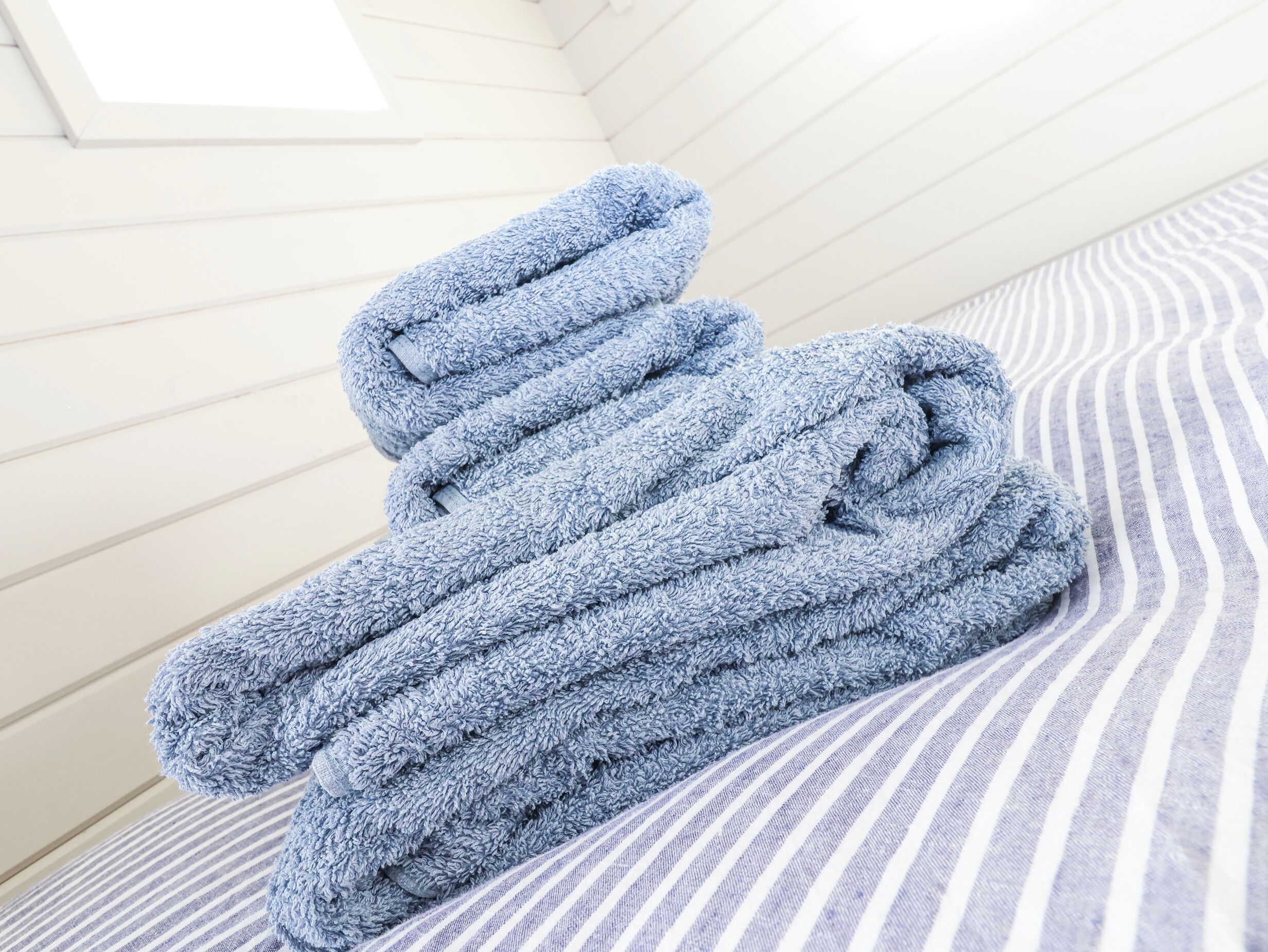 Una toalla azul doblada cuidadosamente sobre una cama a rayas en una habitación con paredes blancas y techo inclinado.
