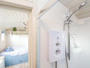 Un baño moderno con ducha, inodoro y lavamanos. Se observa una cama con cubrecama azul y blanco a rayas y una planta pequeña en la repisa.