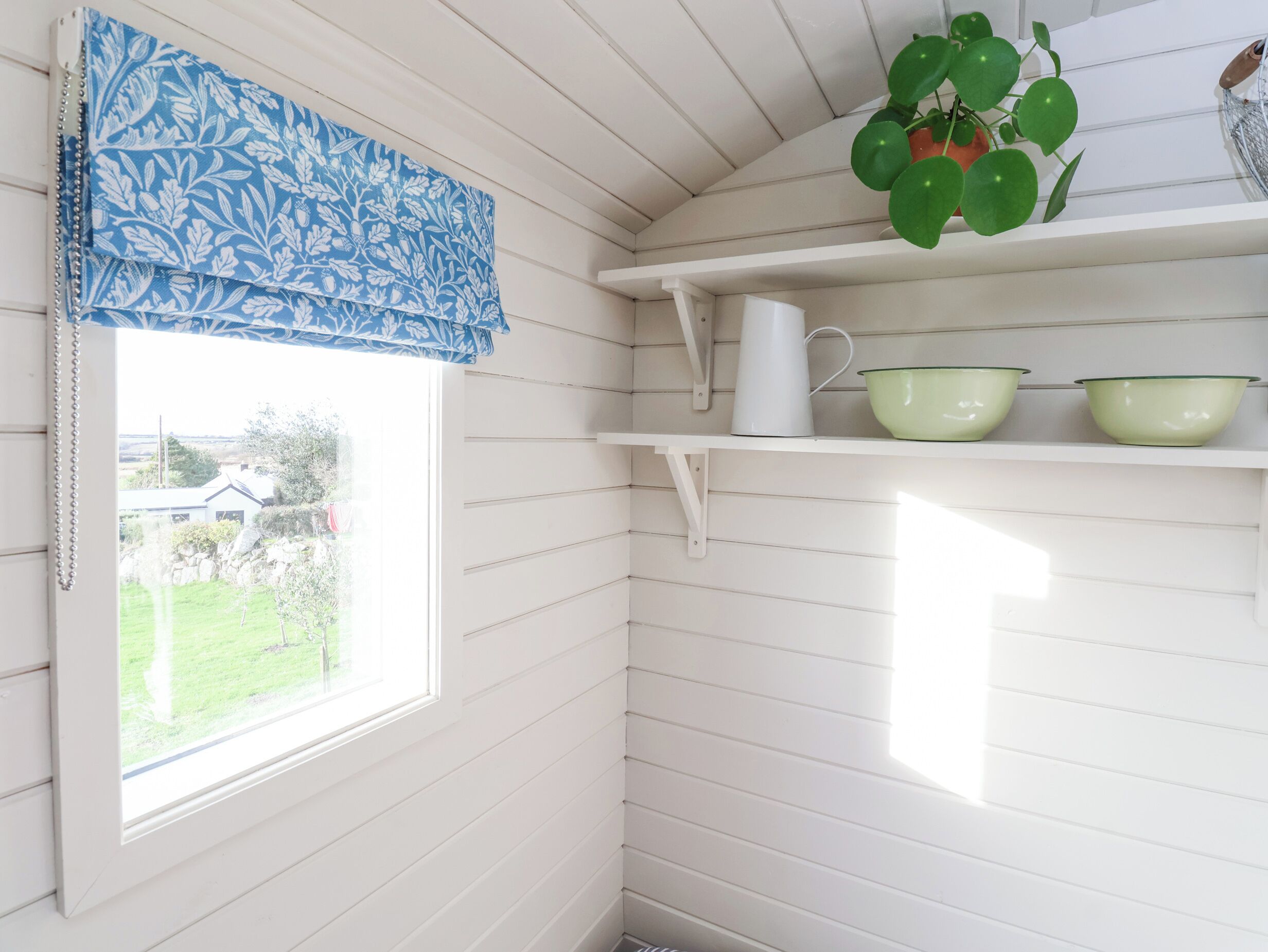 A corner of a room with a window, a blue patterned valance, a white pitcher, and green bowls on a shelf.