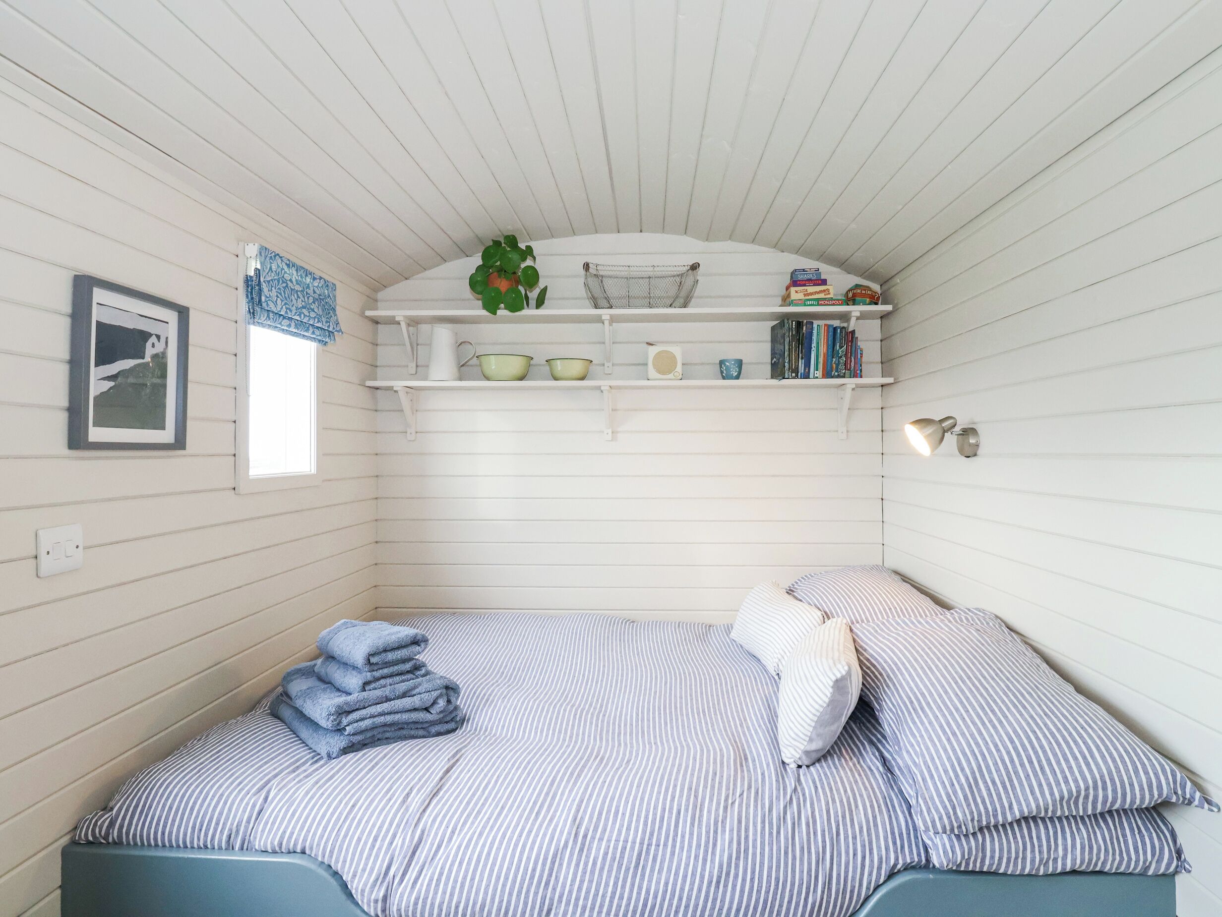 Habitación compacta con una cama individual, un pequeño estante con libros y adornos, un cuadro enmarcado en la pared y una ventana con cortina.