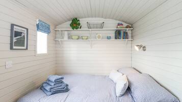 Habitación compacta con una cama individual, un pequeño estante con libros y adornos, un cuadro enmarcado en la pared y una ventana con cortina.