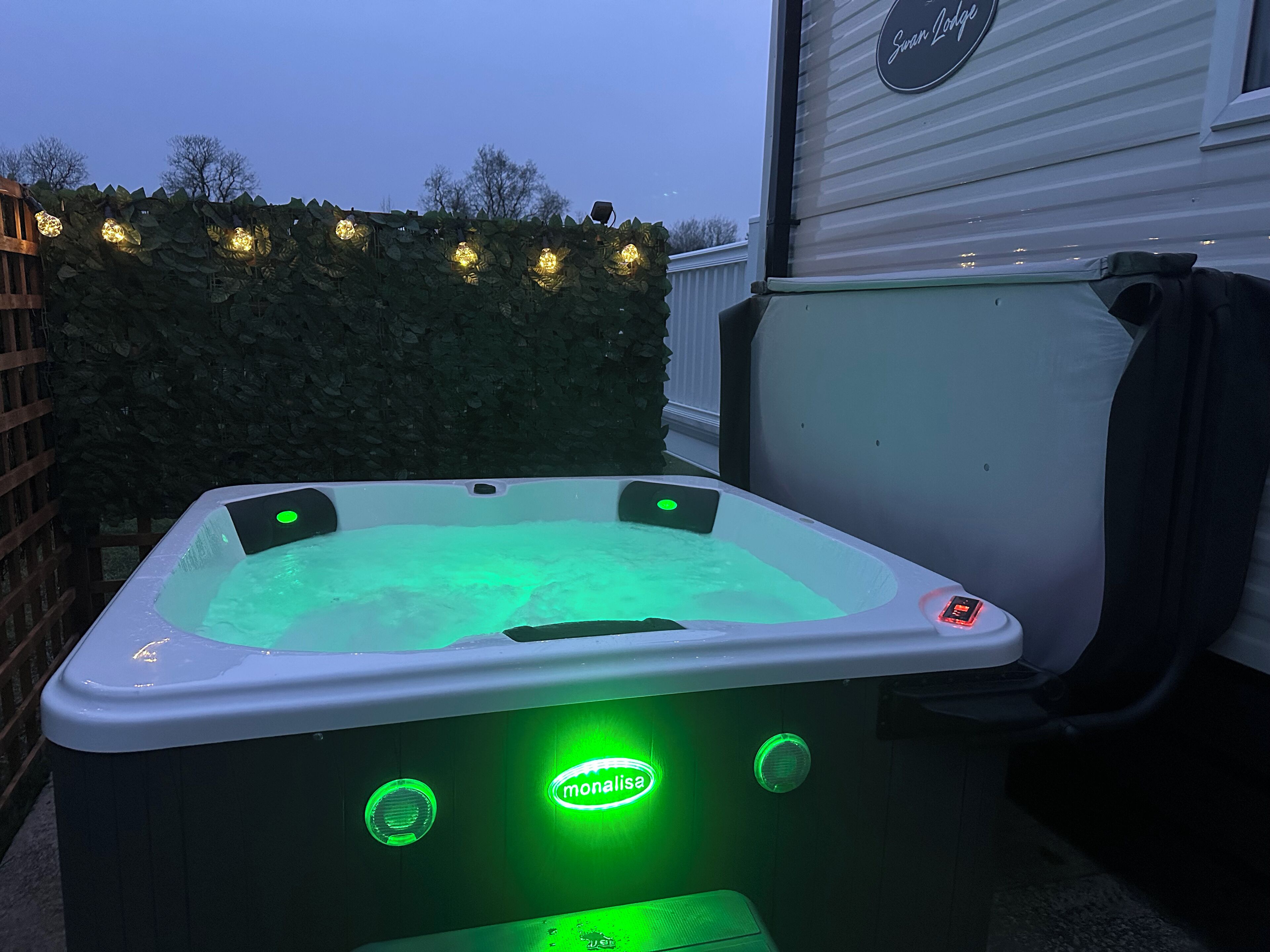 A hot tub with a green light, surrounded by a hedge and string lights.