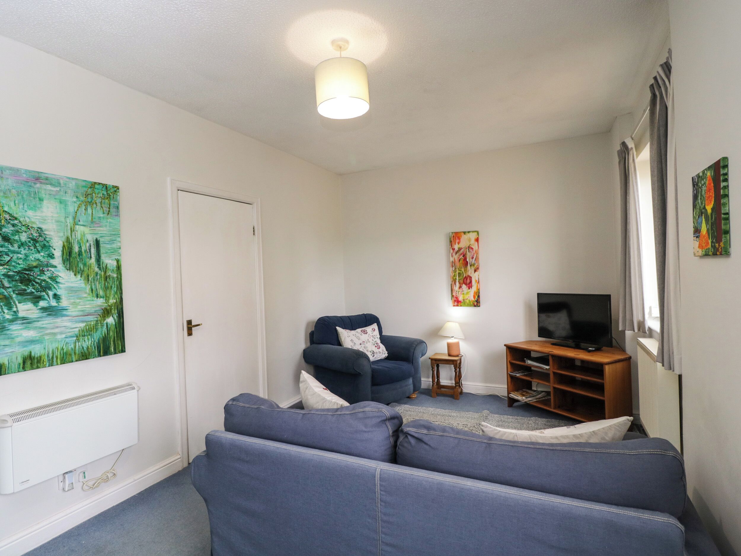 A living room with a blue sofa, a blue armchair, a small wooden side table with a lamp, a television on a wooden stand, and two paintings on the walls.