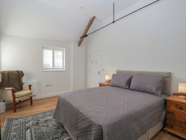 Um quarto com uma cama grande, uma cadeira, uma mesa de cabeceira, teto com vigas de madeira, janela com persianas e um tapete estampado.