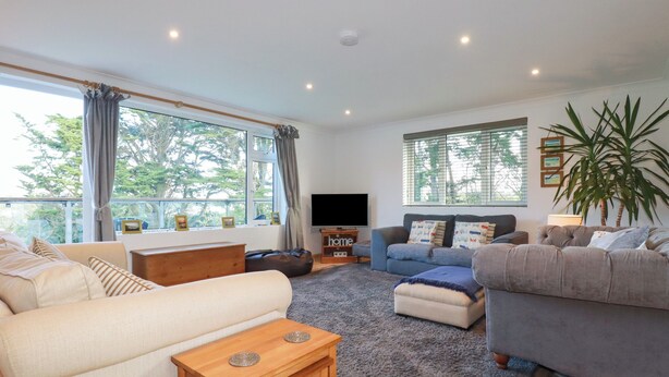A spacious living room with a sofa, armchairs, a coffee table, and a TV. Large windows offer views of trees and a balcony.