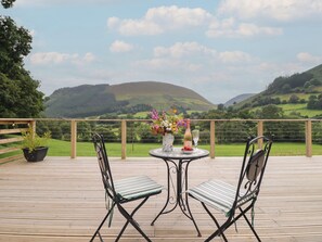 A wooden deck with two chairs and a table set for two, overlooking a green landscape with hills. - Caban Tirion (Machynlleth)