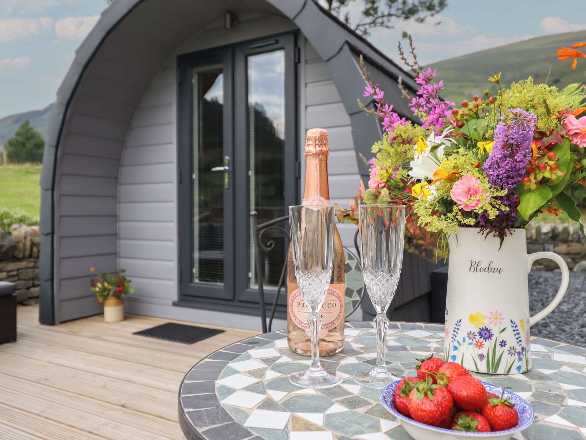 A table with a bottle of Prosecco, two glasses, a bowl of strawberries, and a floral vase.