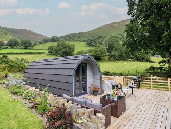 A cozy, modern outdoor seating area with a curved roof structure, surrounded by a wooden deck and stone walls, overlooking a lush green landscape with sheep grazing. - Caban Tirion (Machynlleth)
