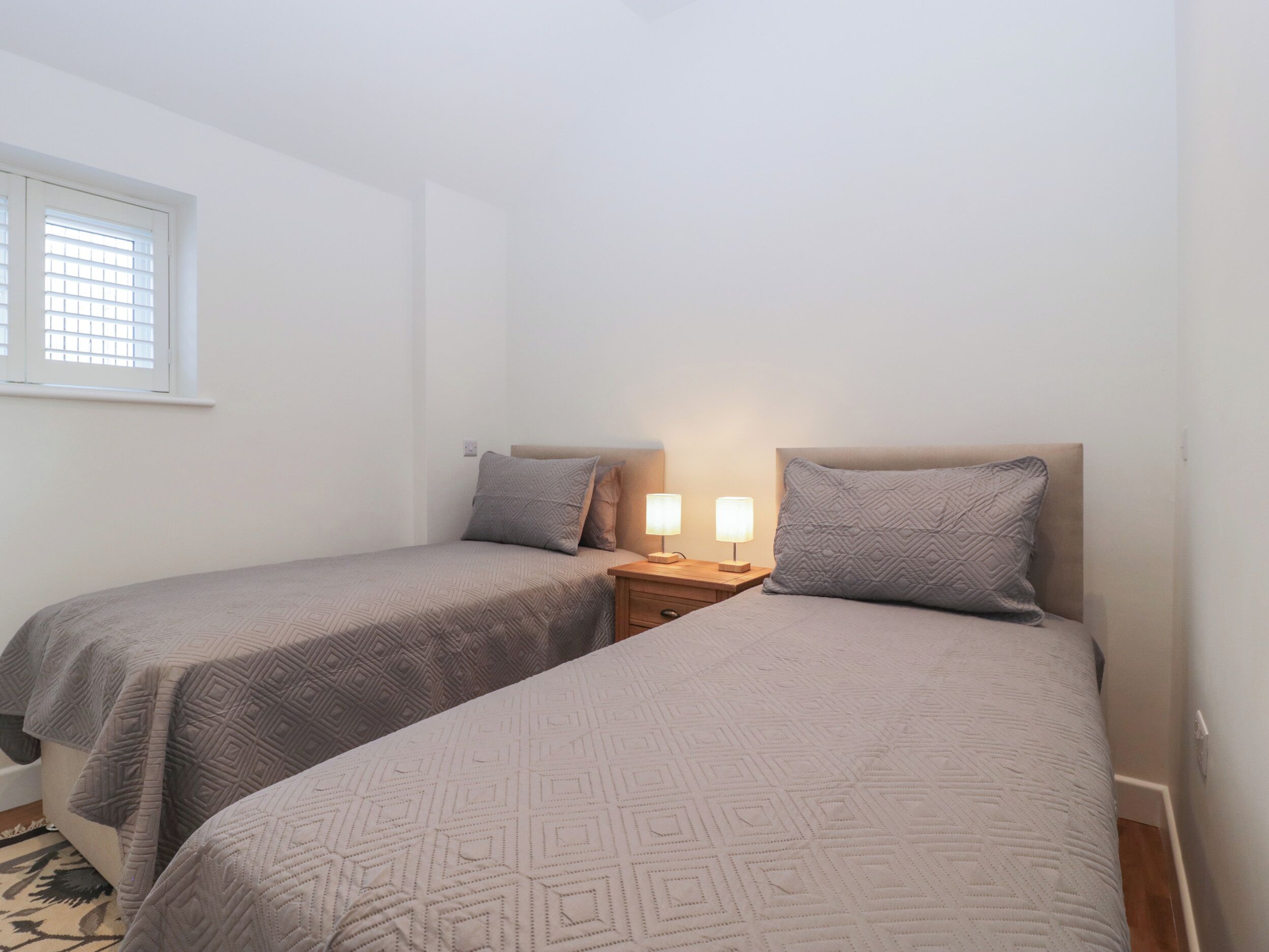 Ein Schlafzimmer mit zwei Einzelbetten, jeweils mit Kopfteil, einem Nachttisch mit Lampe und einem Fenster mit Jalousien.