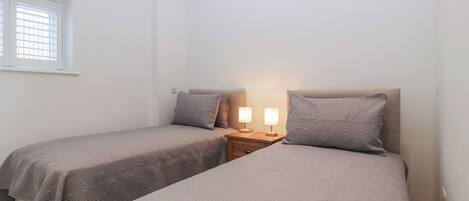 Ein Schlafzimmer mit zwei Einzelbetten, jeweils mit Kopfteil, einem Nachttisch mit Lampe und einem Fenster mit Jalousien.