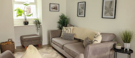 A living room with a grey sofa, a small ottoman, and a black side table with a plant.